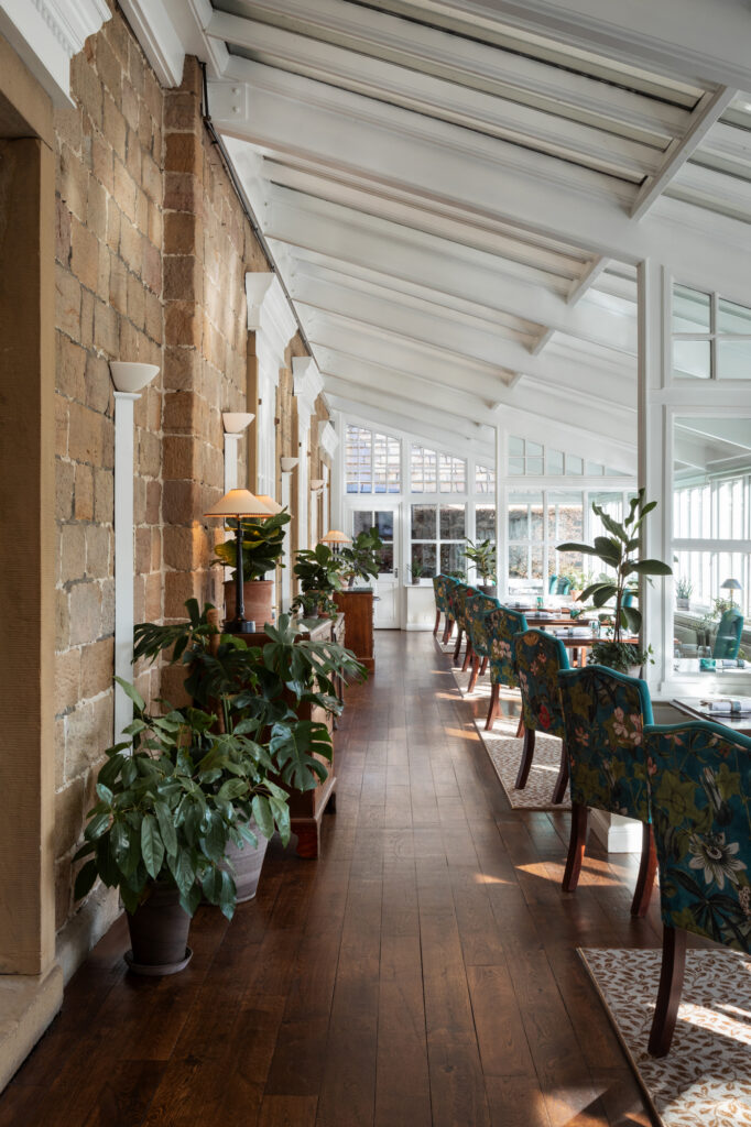 Tables with green floral chairs are surrounded by plants in a large an open conservatory.