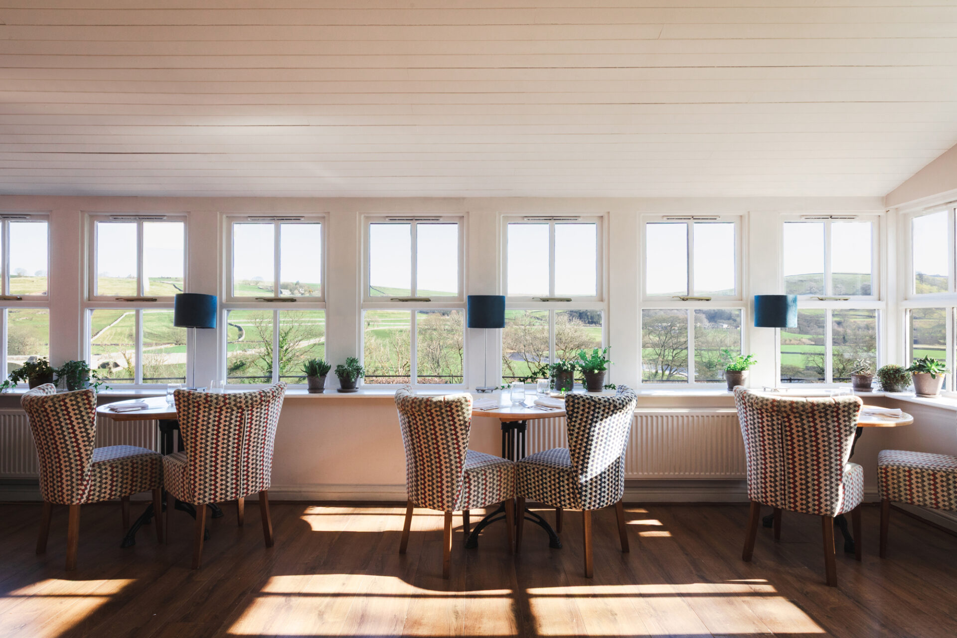 A few tables sit in the large windows of the conservatory with the sunlight streaming through and the view in the background.
