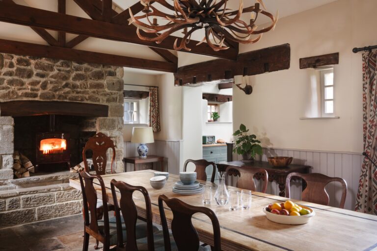 Rustic dining area with long table and roaring open fire with kitchen in the background.