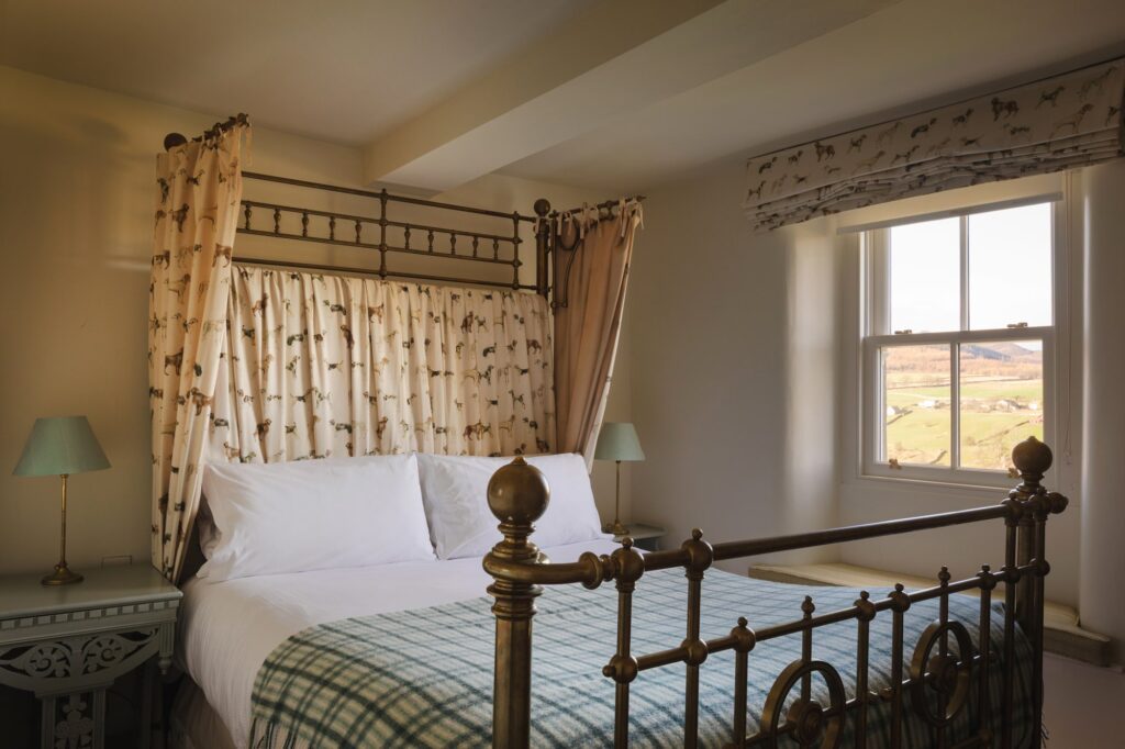 Fourposter bed with dog patterned curtains and check cover, with view of the valley from the window.