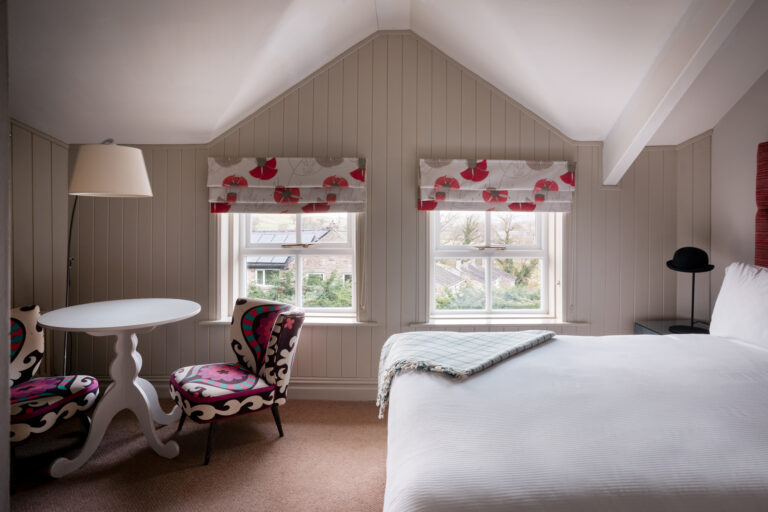 Beige bedroom with wood-paneled walls, floral red blinds, funky patterned chairs around a table and standing lamp.