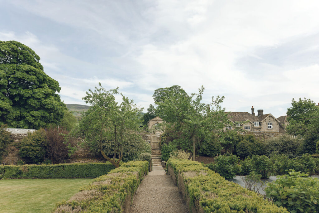 A kitchen garden with lush green bushes and trees with the hotel in the background, there is a little stream visible.