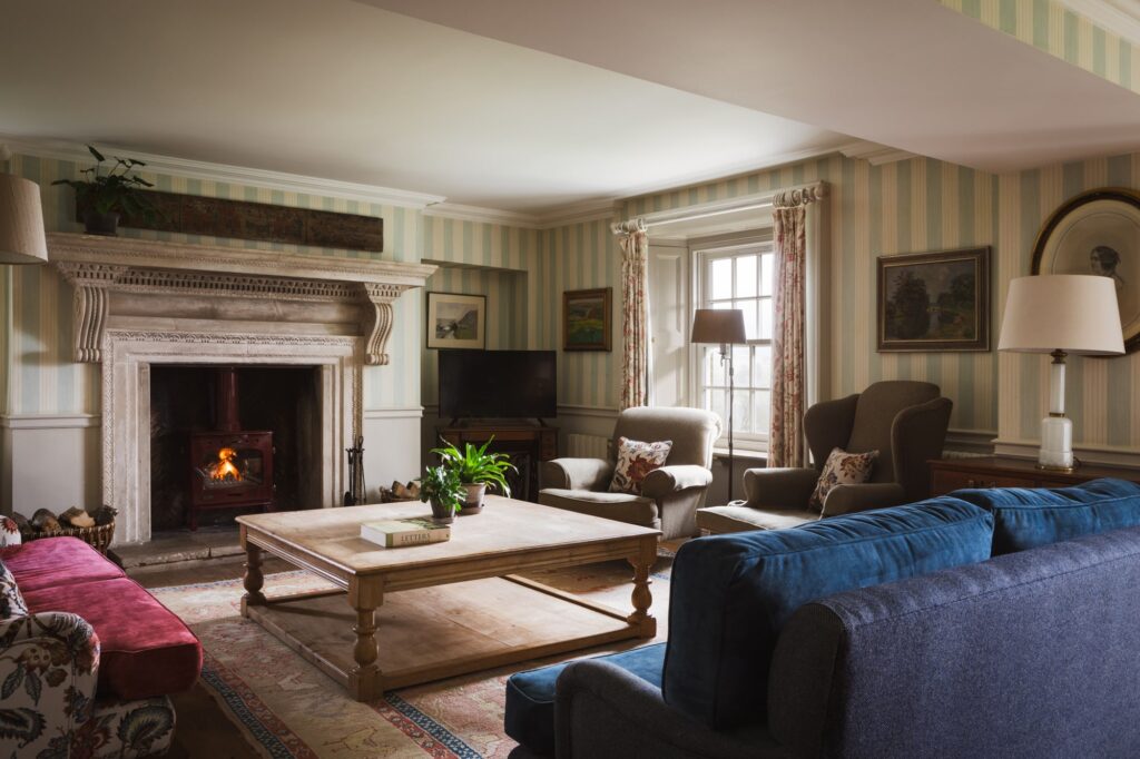 Living area with striped wallpaper and roaring open fireplace, antique furnishings and charm.
