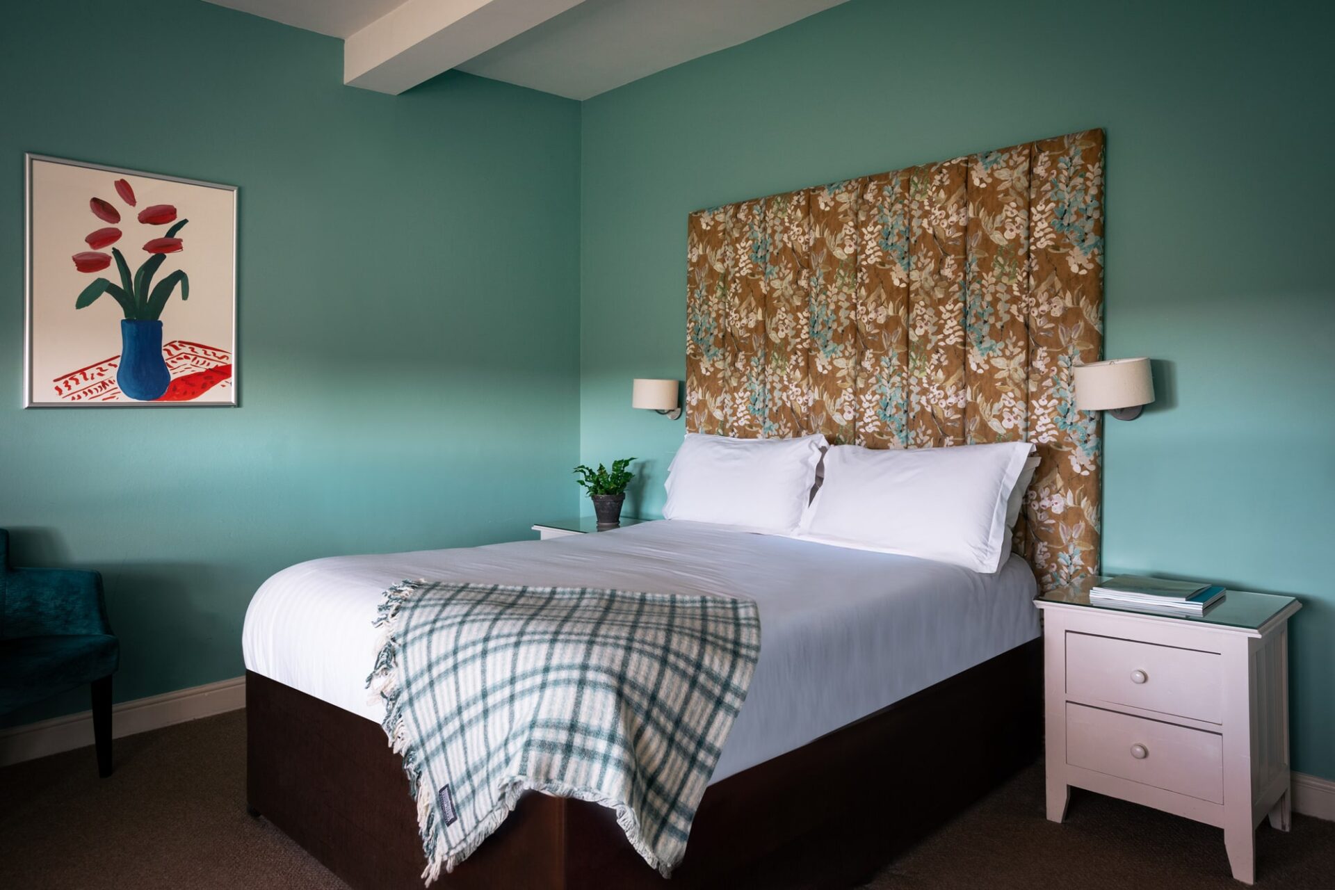 A blue bedroom with yellow and blue floral headboard and check throw with floral artwork.