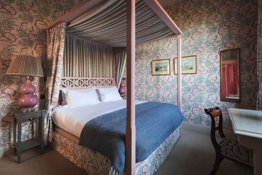 A pink four-poster bed with big floral wallpaper in a cosy room.