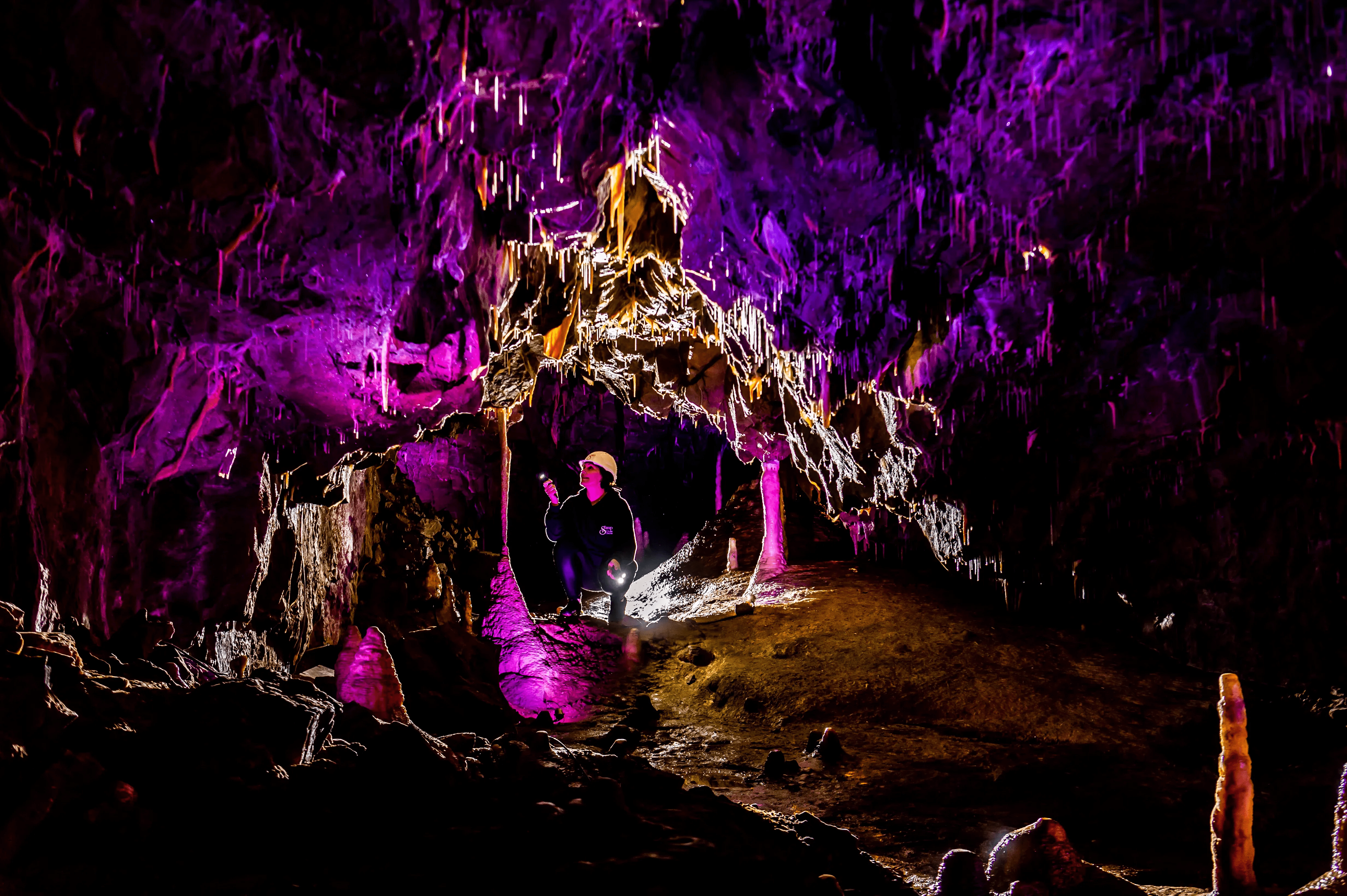An ancient cave is aglow with purple lights, a woman crouches to look inside a hole.