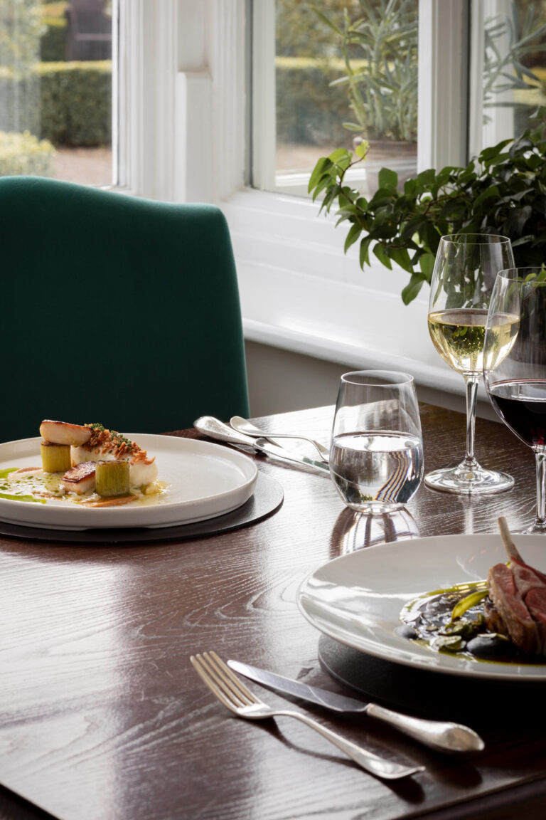 A table of food with cutlery and napkins, white and red wine and a green chair.