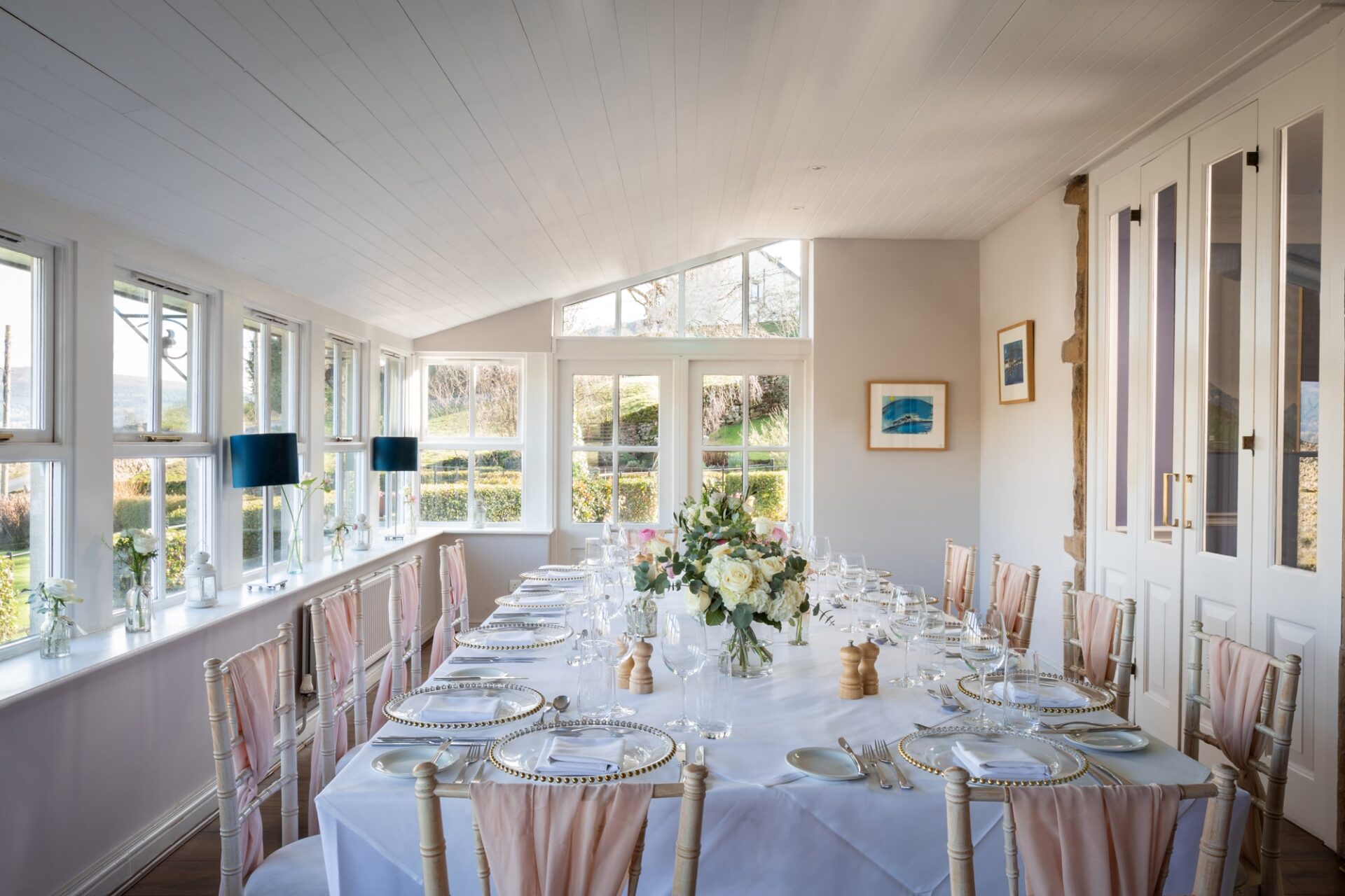 A large private dining table is set up in the conservatory with pink sashes on the chairs and windows looking out onto the view and garden.