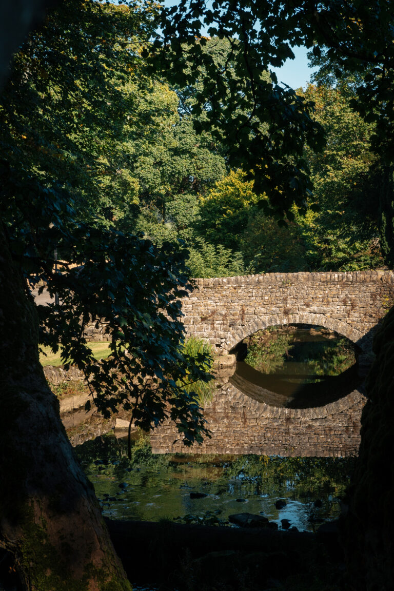 A small stream is running underneath an old stone bridge with the trees surrounding it.