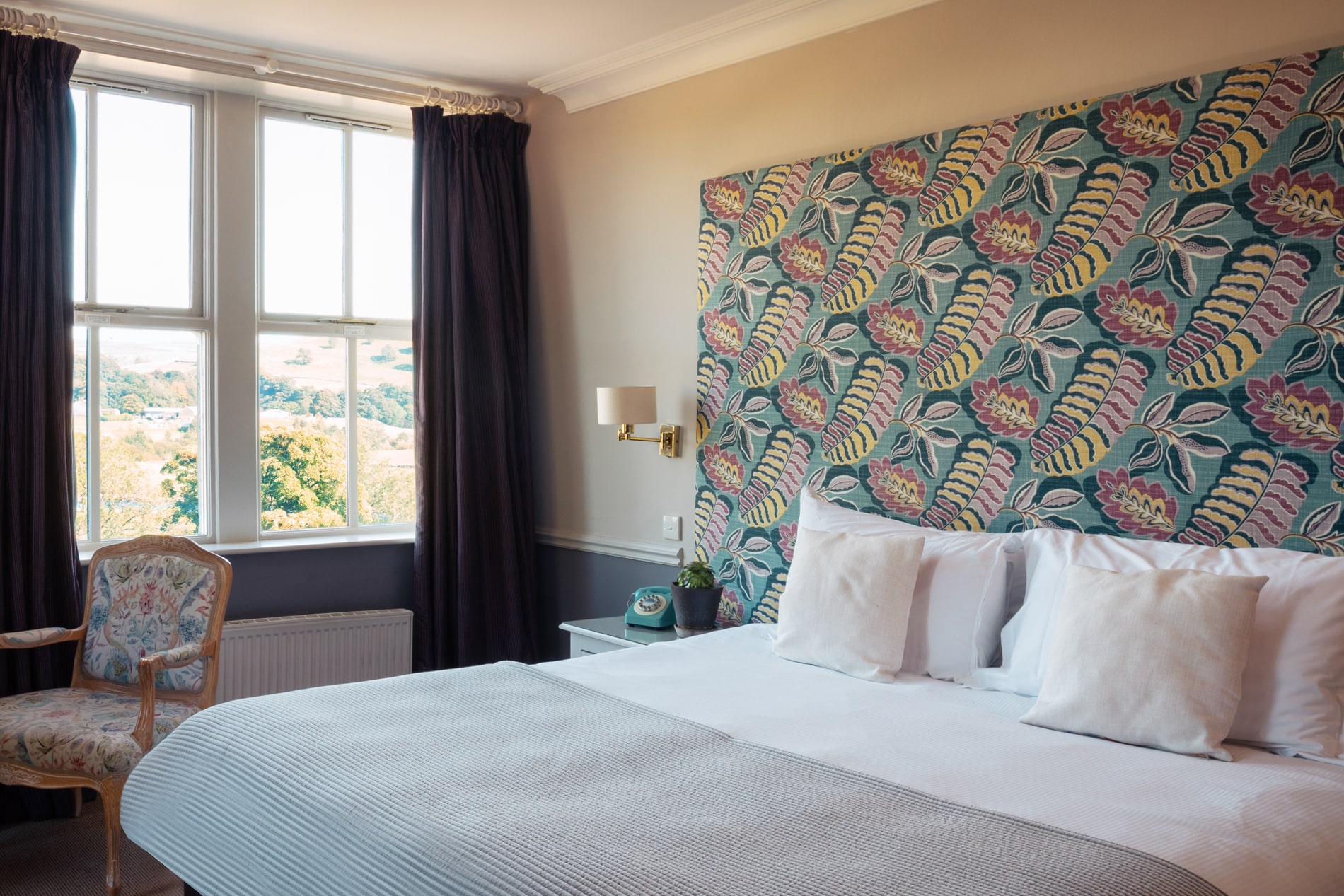 A blue floral headboard in a large room with purple curtains and floral chair.