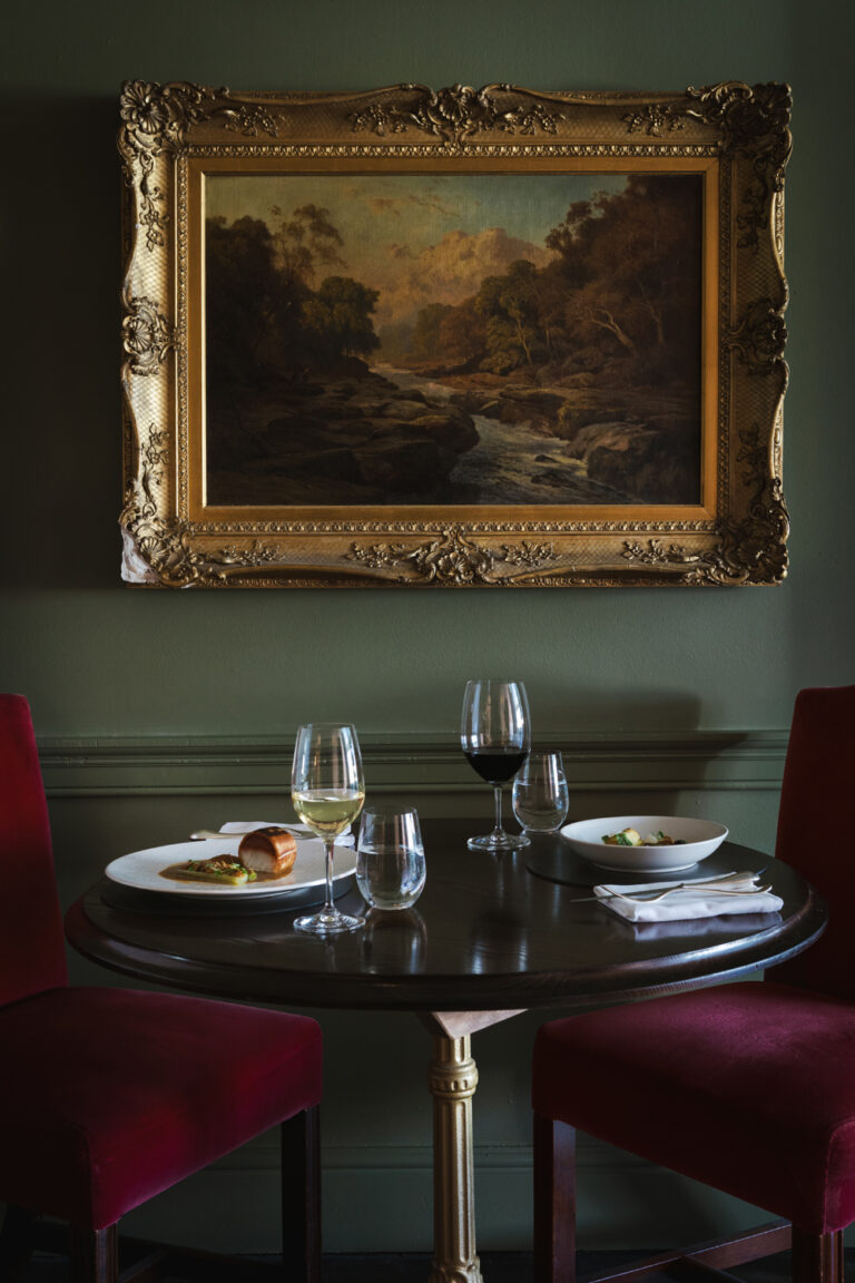 A table is set with food and wine in a green painted room with oil painting behind.