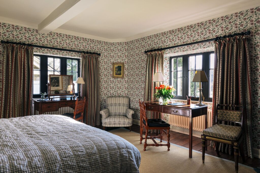 A bedroom with floral wallpaper and a blue bed, the room has two old oak desks and chairs in it.