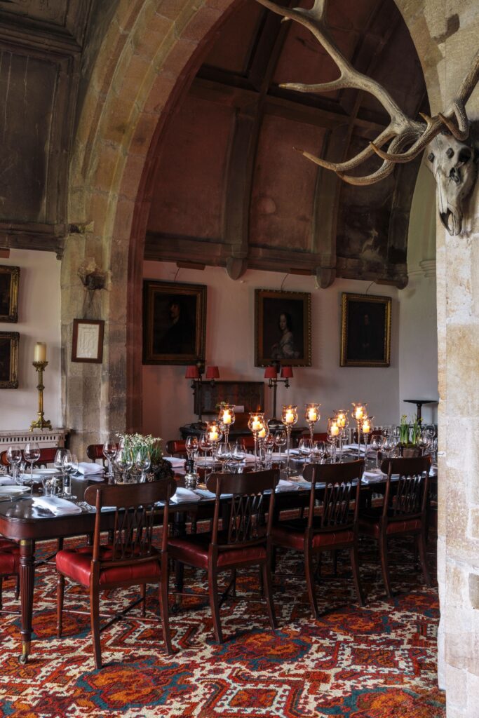 A large oak dining table with candles on surrounded by oil paintings in the arch of a stone hall.