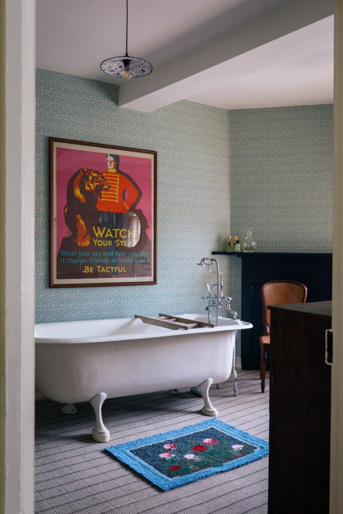 A bathroom with blue wallpaper and a large poster over a roll top bath with antique furnishings.