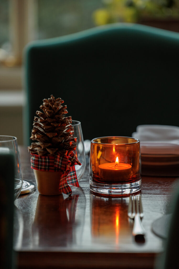 A table set for dinner, with a green chair behind. A amber lit tea light sits on the table next to a small pine cone as decoration.