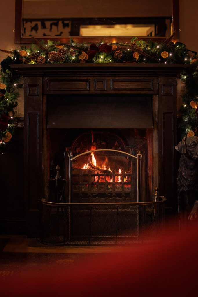 A large fire place, with a roaring fire. A Christmas garland lays above.