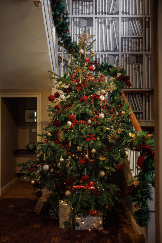 A large Christmas tree with red baubles, tinsel and crackers laid on it. Gold and white wrapped presents lay beneath, by a staircase.