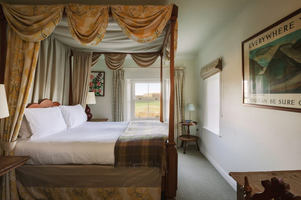 Large bedroom with fourposter bed, with yellow draped curtains and window with valley view.
