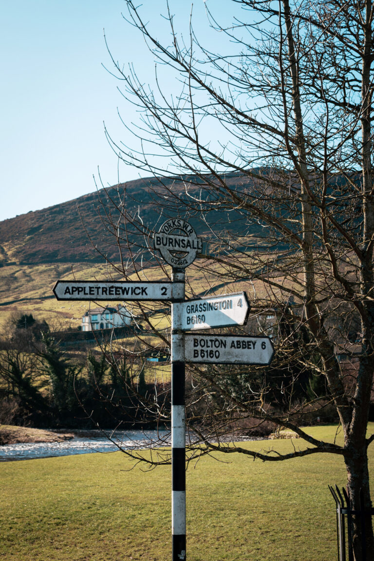 An old fashioned signpost for the Yorkshire Dales, with nearby town names with black and white stripes and the rolling hills beyond.
