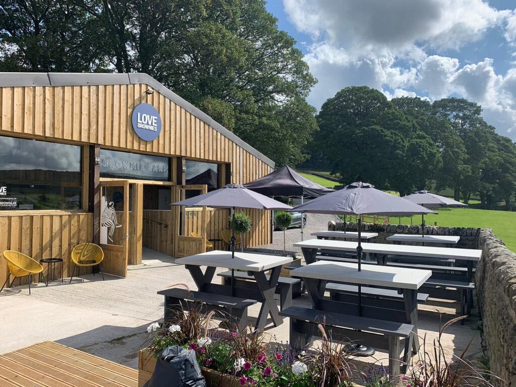 The wooden barn is in the sunlight with benches and umbrellas outside.