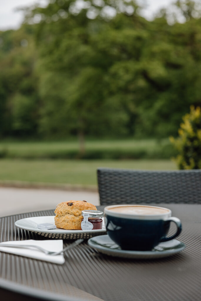 A scone with jam and butter with a coffee placed on a table overlooking green scenery.