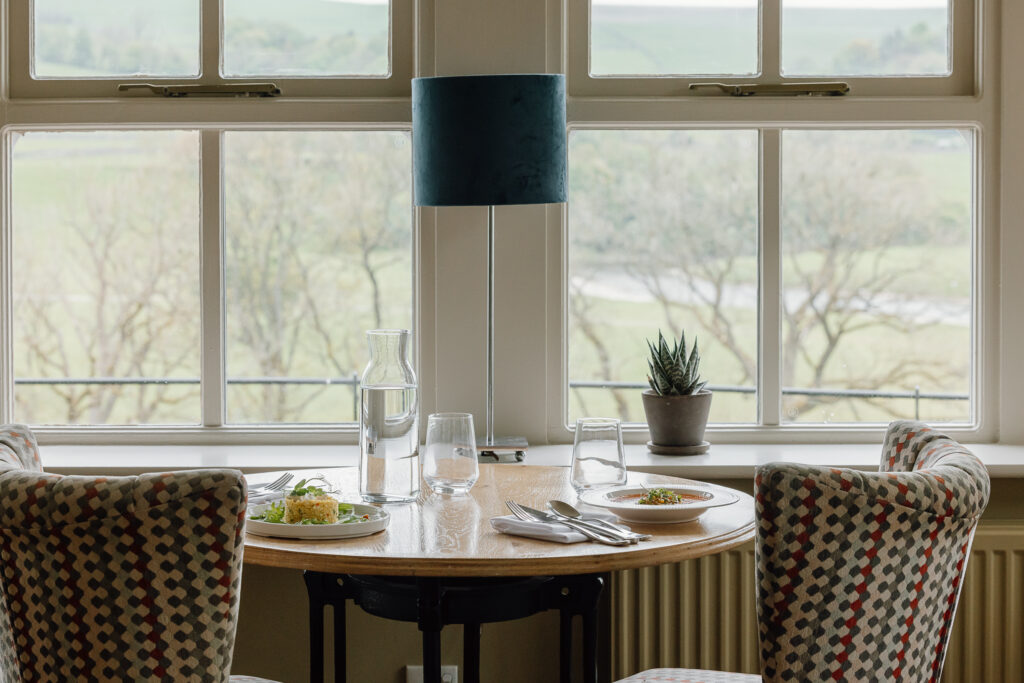 Two summer dishes of food sat on a table, there is two checked chairs behind overlooking the yorkshire dales and river.